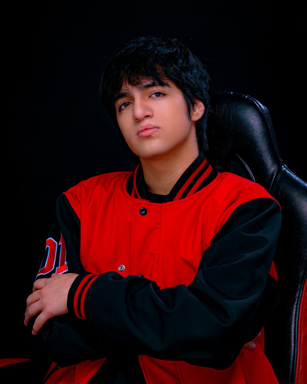 Evans The Champion con los brazos cruzados en su silla gamer, con chaqueta varsity roja.