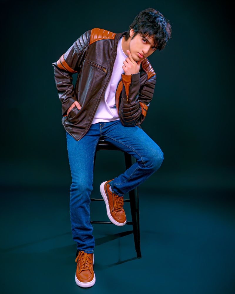 sentado en un taburete, con chaqueta de cuero marrón, jeans y zapatillas